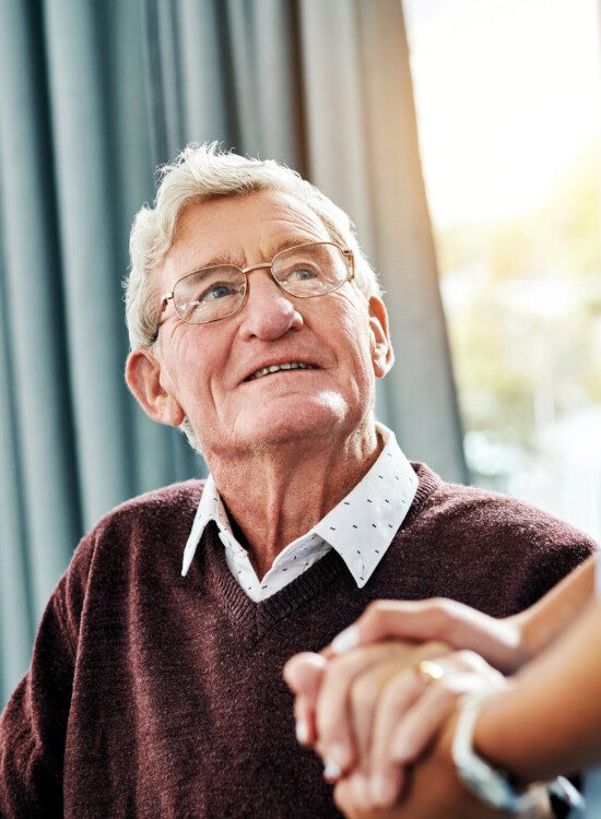 senior man smiles up at caregiver while seated, holding her hand