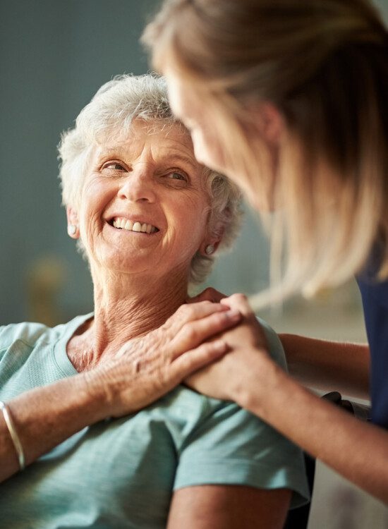 senior woman smiles up at caregiver, who gently places her hands on her shoulder