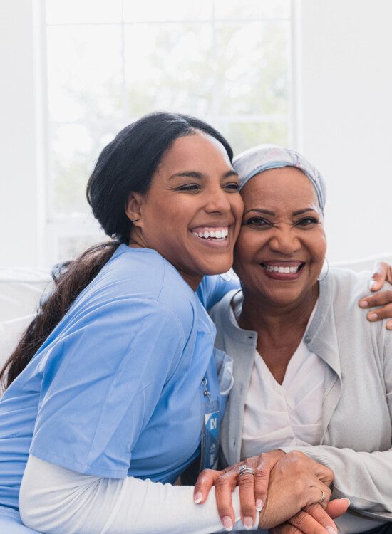 senior woman and her caregiver smile and hug while seated on a couch
