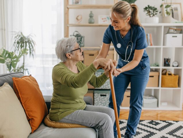 seated senior woman with cane smiles up at her female caregiver, who reaches down to help her stand