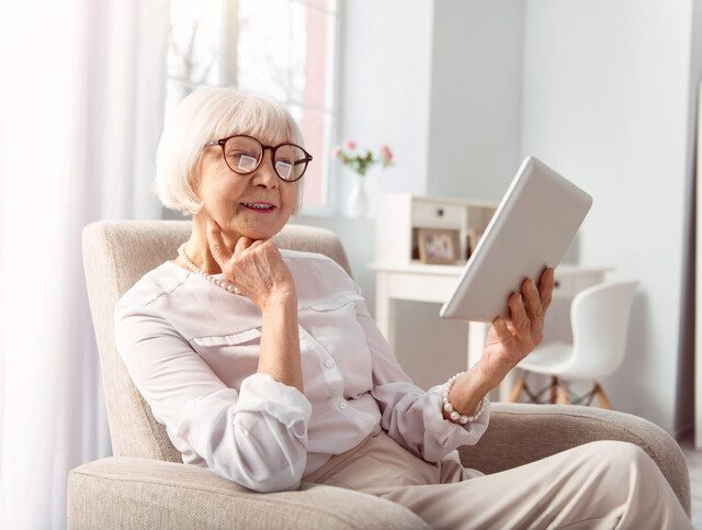 elegant senior woman sits in a reclining chair in her apartment, reading while resting her head on her hand