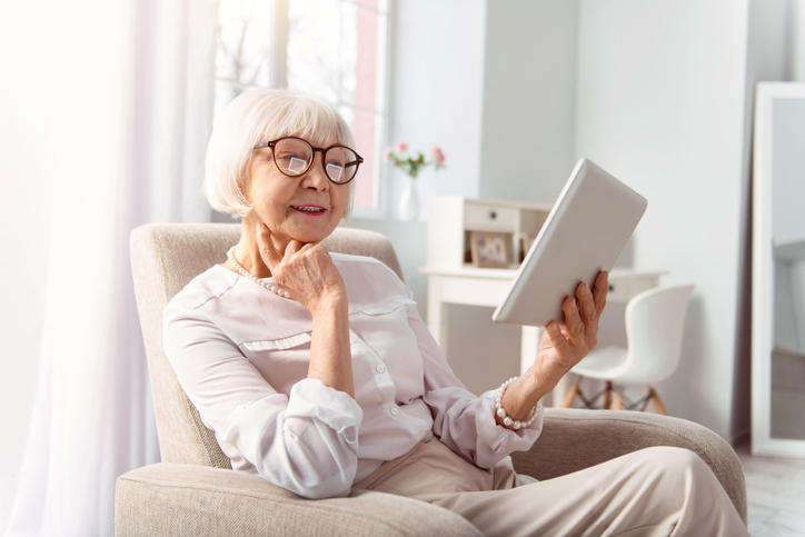 elegant senior woman sits in a reclining chair in her apartment, reading while resting her head on her hand
