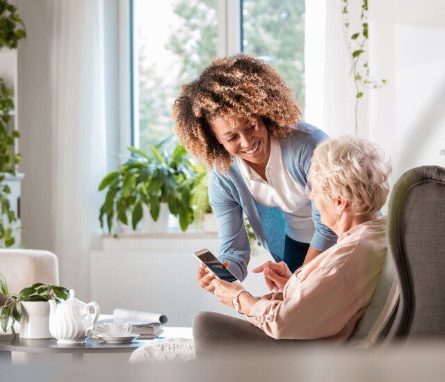 seated senior woman smiles up at her female caregiver, who's helping her navigate her smart phone