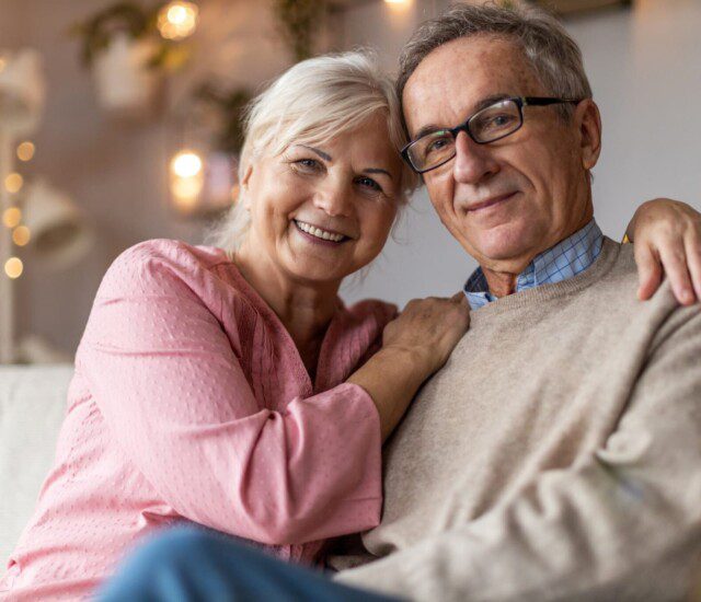 senior woman smiles while snuggling her husband on the couch, both looking at the camera