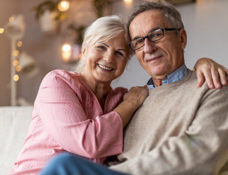 senior woman smiles while snuggling her husband on the couch, both looking at the camera