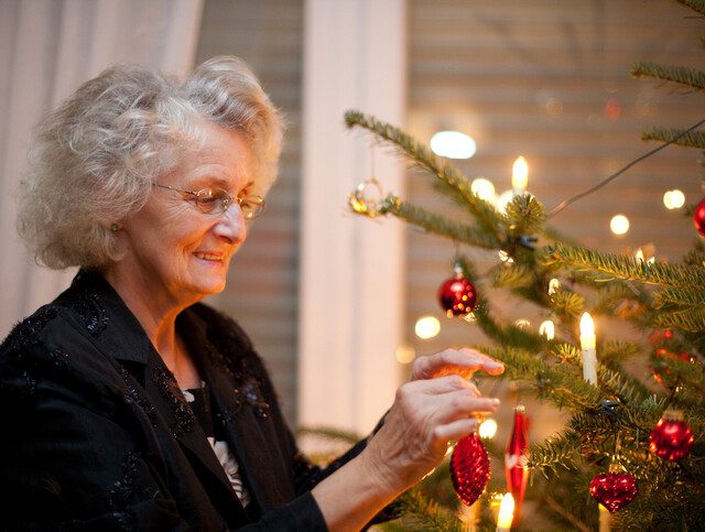 senior woman smiles while gently placing a red Christmas ornament on a tree