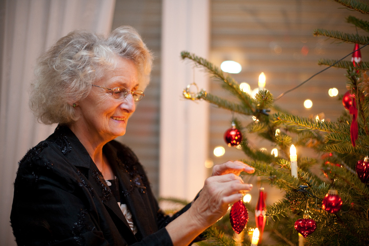 senior woman smiles while gently placing a red Christmas ornament on a tree