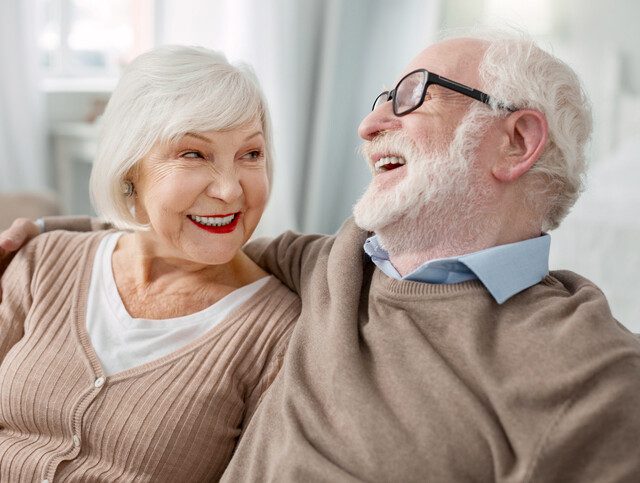 senior woman looks lovingly at her husband while he laughs, both seated on a couch snuggling