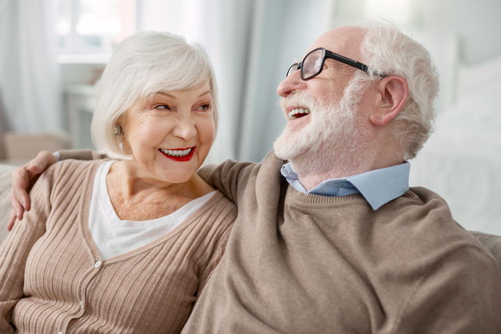 senior woman looks lovingly at her husband while he laughs, both seated on a couch snuggling