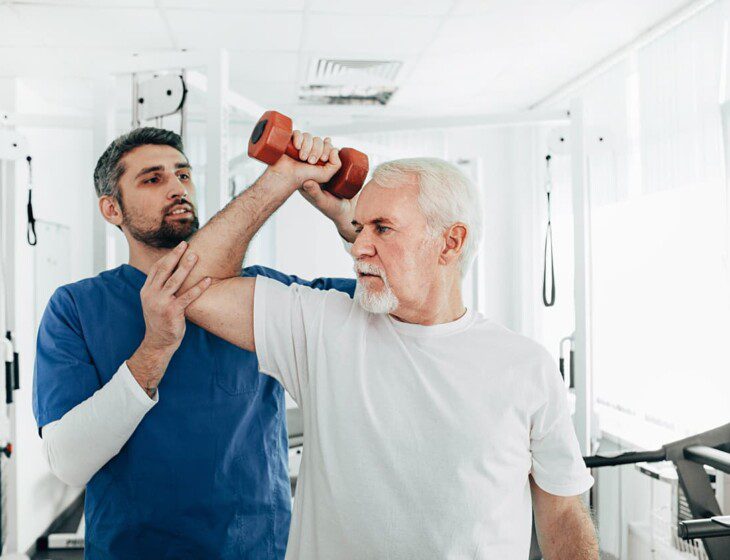 senior man holds dumbbell in one arm with the help of occupational therapist