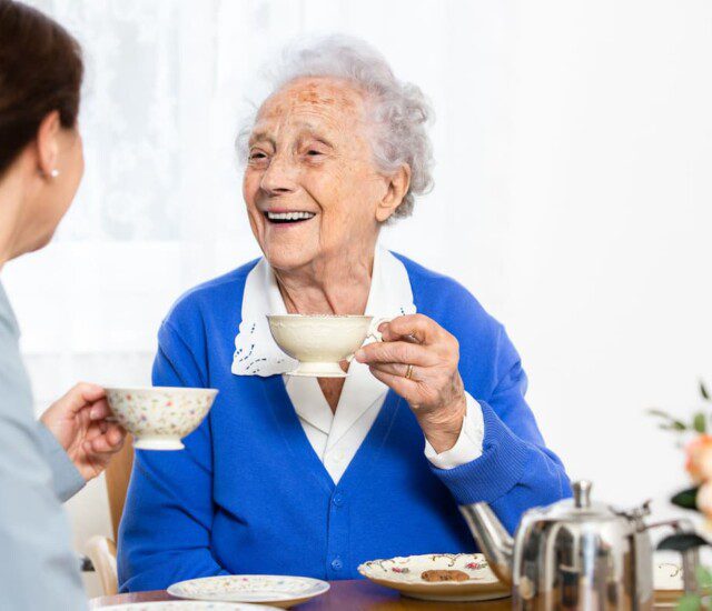 two senior woman sit at a table and drink tea, smiling at one another