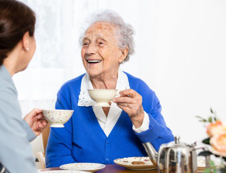 two senior woman sit at a table and drink tea, smiling at one another