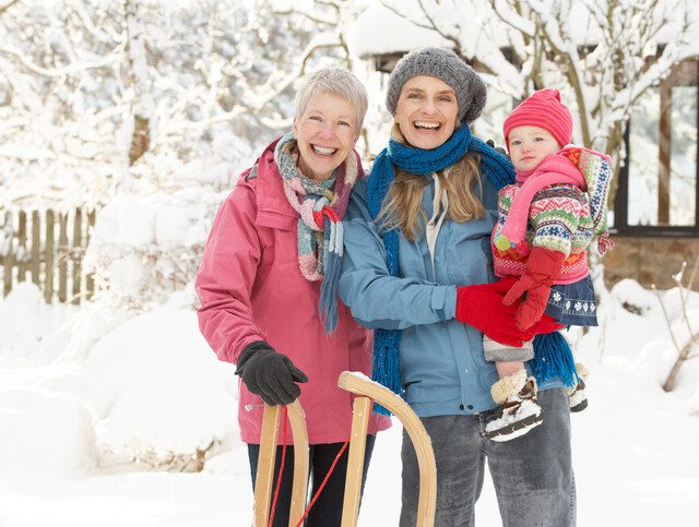 senior woman, her adult daughter, and her granddaughter smile at the camera, buddled up for sledding