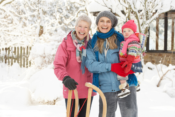 senior woman, her adult daughter, and her granddaughter smile at the camera, buddled up for sledding
