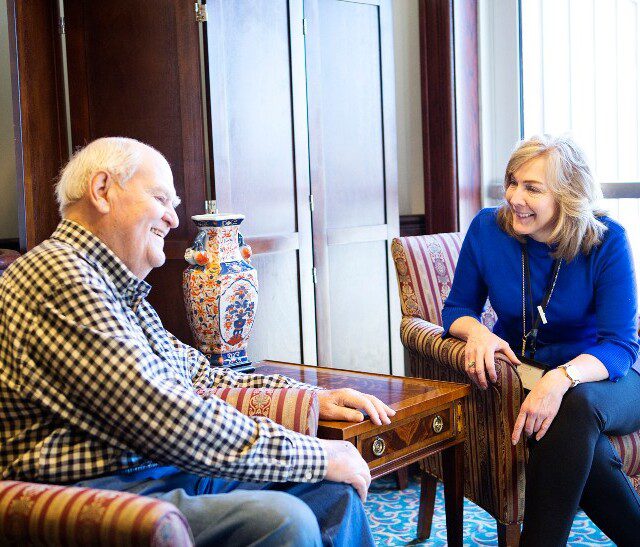 male and female senior friends smile and converse, seated in chairs next to one another in a lounge