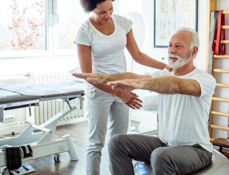 senior man works on mobility while seated on an exercise ball, guided by a physical therapist