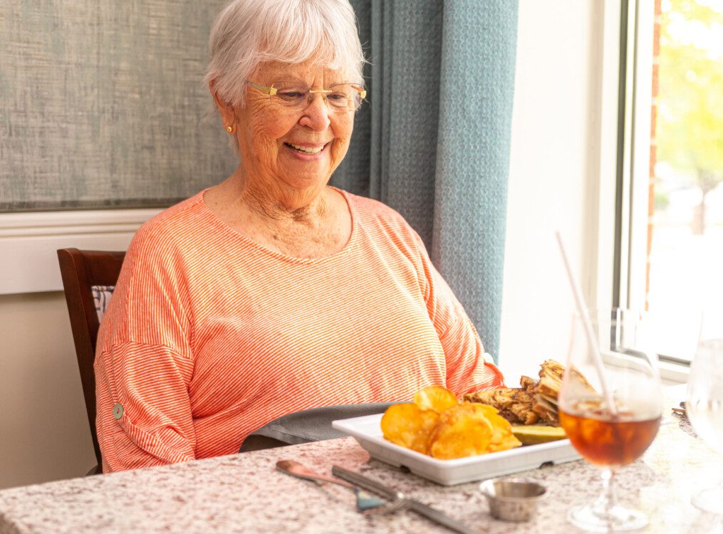 Seated senior woman smiles down at the plate of delicious food before her on the table