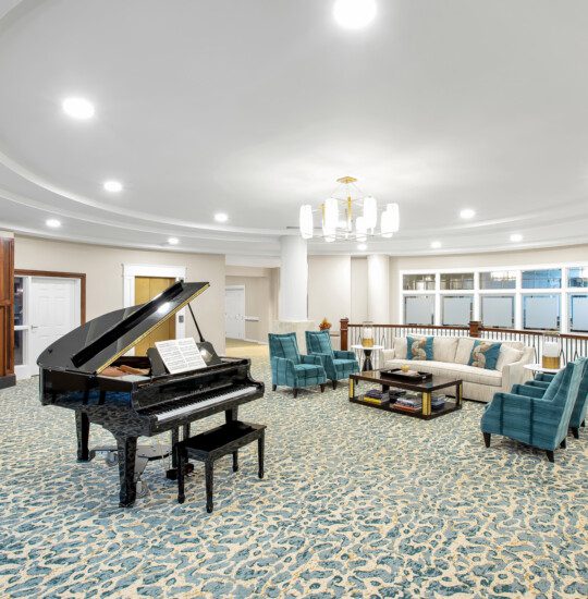 elegant second-floor lounge with seating, fireplace, and grand piano at Claridge Court Senior Living Community