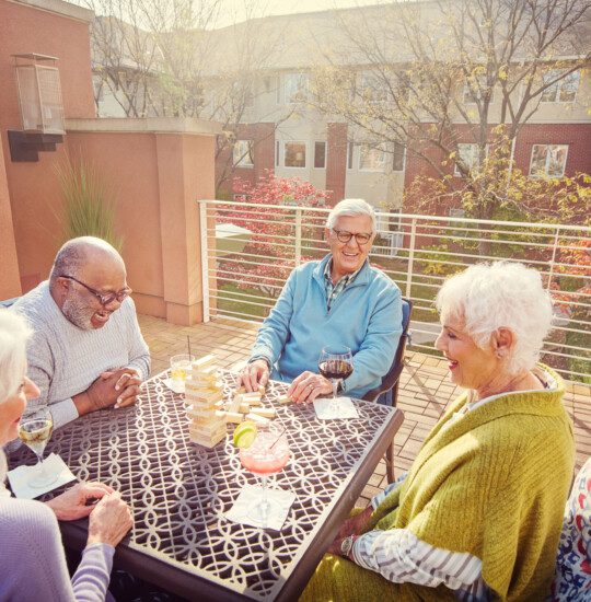 group of senior friends sit and play cards while drinking cocktails on the scenic outdoor terrace at Claridge Court Senior Living Community