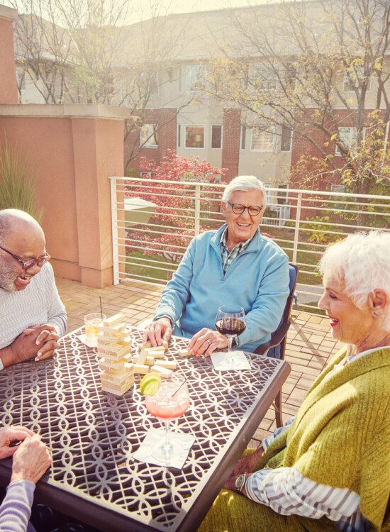 group of senior friends sit and play cards while drinking cocktails on the scenic outdoor terrace at Claridge Court Senior Living Community