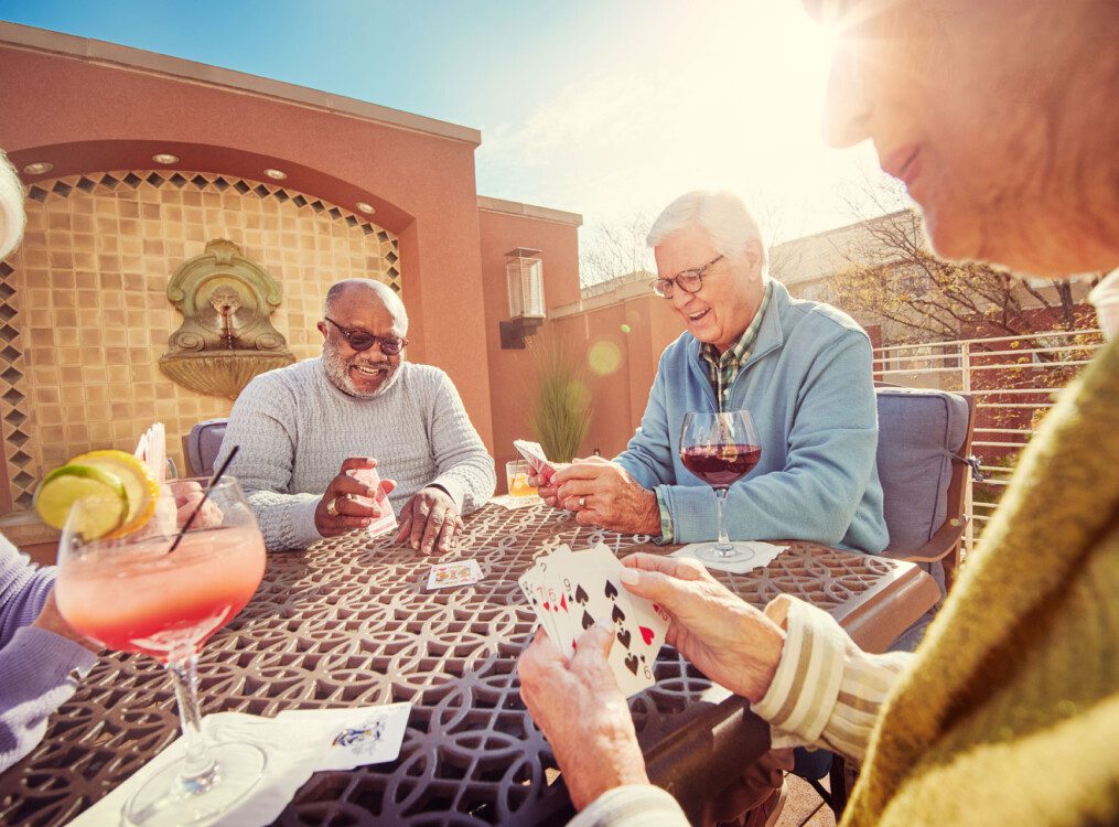 group of senior friends sit and play cards while drinking cocktails on the scenic outdoor terrace at Claridge Court Senior Living Community