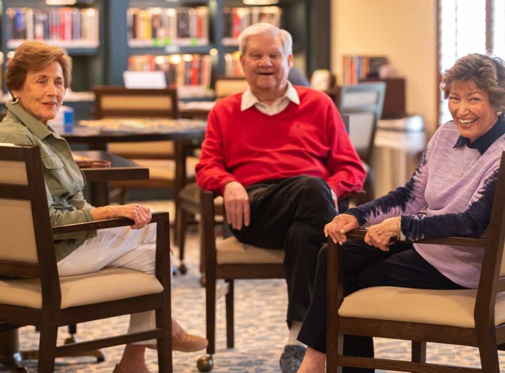 three seniors in chairs in the library at Claridge Court turn to face the camera and smile