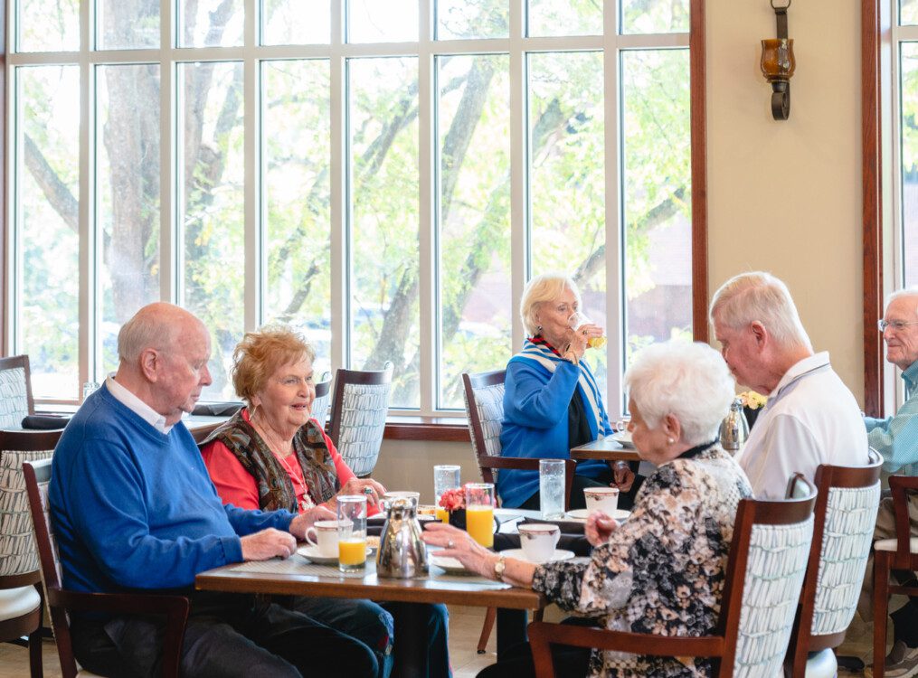 group of seniors smile and sit at a table in a casual dining space for brunch together at Claridge Court Senior Living Community