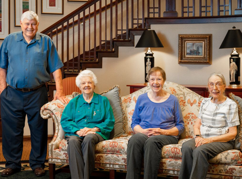 three female seniors seated on a couch and one standing senior male resting a hand on the couch, all smiling for an interview