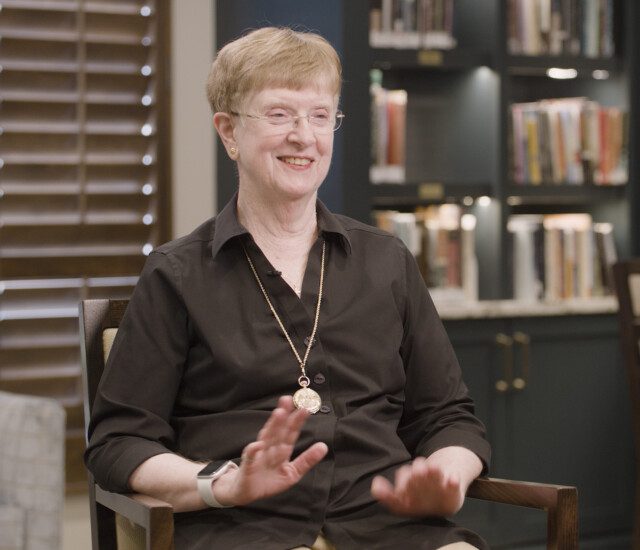 Senior woman in black button-up shirt smiles and sits in chair for interview