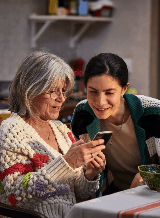 Two women share a smile while looking at a smartphone together indoors.