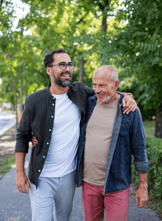 Two men walk together on a sunny day with trees and greenery around them.