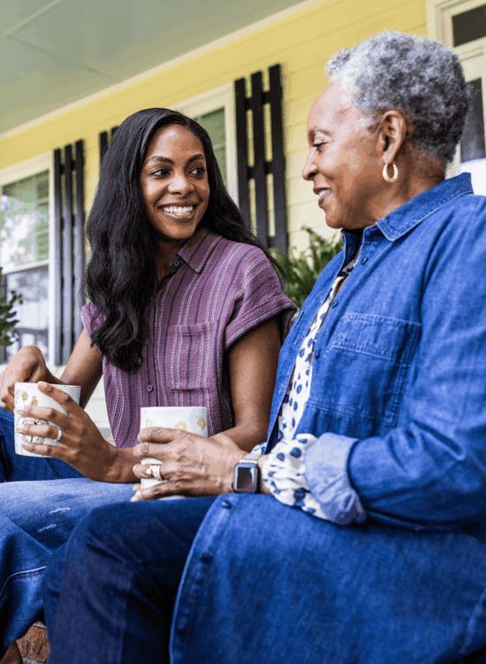 Two women share smiles and coffee on porch steps.