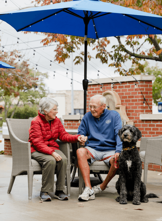 Elderly couple and dog share a moment under an umbrella on patio.
