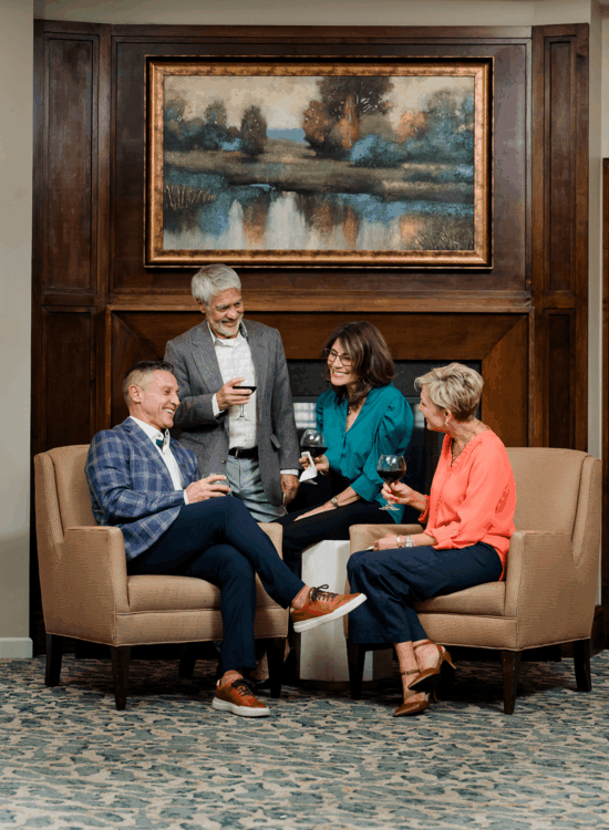 Four people enjoy drinks and conversation in an elegant room with artwork on wood-paneled wall.