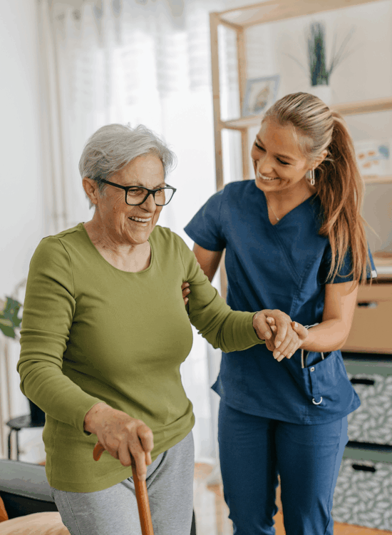 A caregiver assists an elderly woman with walking using a cane indoors.